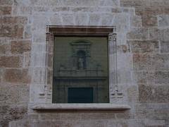 Window of Palau de Benicarló reflecting the entrance of Sant Llorenç church in Valencia