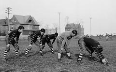 Five Hyde Park High School football players poised to start a play on a Chicago athletic field in 1902 with houses in the background