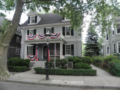 Two story gray house with dormers and red white blue bunting on porch railings with flag flying and gas lamp