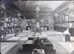 Interior view of Galerie Vasnier room with skylight ceiling, walls covered with paintings, sculptures on pedestals, art objects in display cases, and rugs on the floor