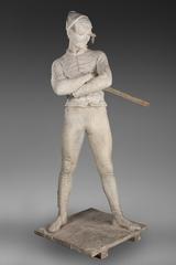 Sculpture of Arlequin standing with legs apart wearing a mask and hat, face tilted to left, arms crossed on chest holding a stick