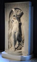 Nude Arab woman wearing babouches standing at the threshold of a Moorish house, lifting a curtain with her right hand, surrounded by geometric, interlace, and stylized floral bas-relief decoration