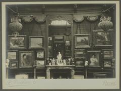 Interior view of Galerie Henry Vasnier cimase with paintings hung on the wall and some resting on the floor, centered around a fireplace topped with a mirror reflecting the photographer. Sculptures and art objects are displayed on the fireplace mantle and on columns.