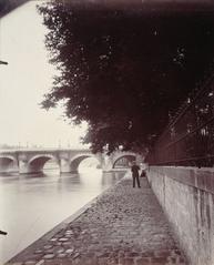 Black and white photograph of the north side banks of Vert-Galant island near Pont Neuf in Paris showing Seine river quay, trees, and a solitary man with the Pont Neuf bridge in the background