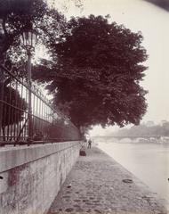 Historical black and white photograph of the northern banks of Vert-Galant island in Paris, showing the Seine quayside with trees, people including a painter at an easel, and the Pont des Arts bridge in the background, taken in 1910.