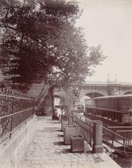 Black and white photograph of Pont Neuf and the Seine quays in Paris with trees, a barrière, bacs, and a péniche, taken in 1910 by Eugène Atget