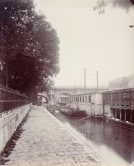 Black and white photograph of the southern bank of Vert-Galant, Seine river with trees, a barge, and the Pont Neuf bridge in Paris