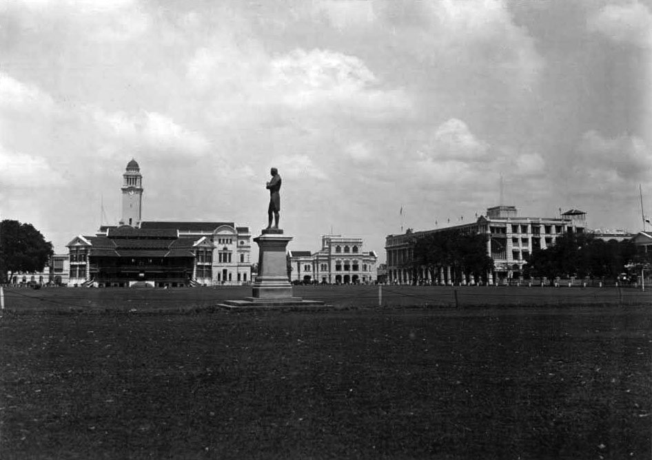 View of the Padang in Singapore with a statue of Sir Stamford Raffles in the foreground and historic buildings including Singapore Cricket Club, Town Hall, Supreme Court, and Grand Hotel de l'Europe i
