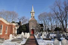 Snow covered burial ground in front of Gloria Dei, a historic brick church with a red door and wooden steeple