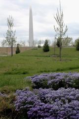 Side view of the Gateway Arch with south leg visible, green grass, purple flowers and sporadic trees on the grounds