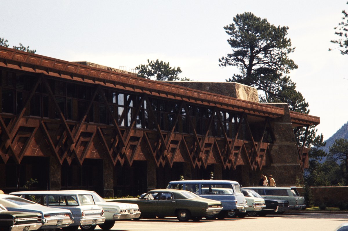 Cars parked outside Mission 66 building surrounded by trees and mountains in Rocky Mountain National Park