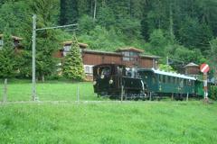 Steam locomotive G 3/3 6 pulling a historic passenger train on the Blonay–Chamby Museum Railway in summer 2010 near Bifurcation station with the museum's vehicle hall in the background