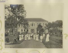 Historical photograph of Cholon municipal hospital staff including Vietnamese and French personnel in front of hospital building with signs for Direction, Economat, Doctors, and Pharmacy