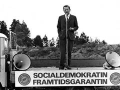Olof Palme giving a speech on a truck in an outdoor setting with pine trees in the background