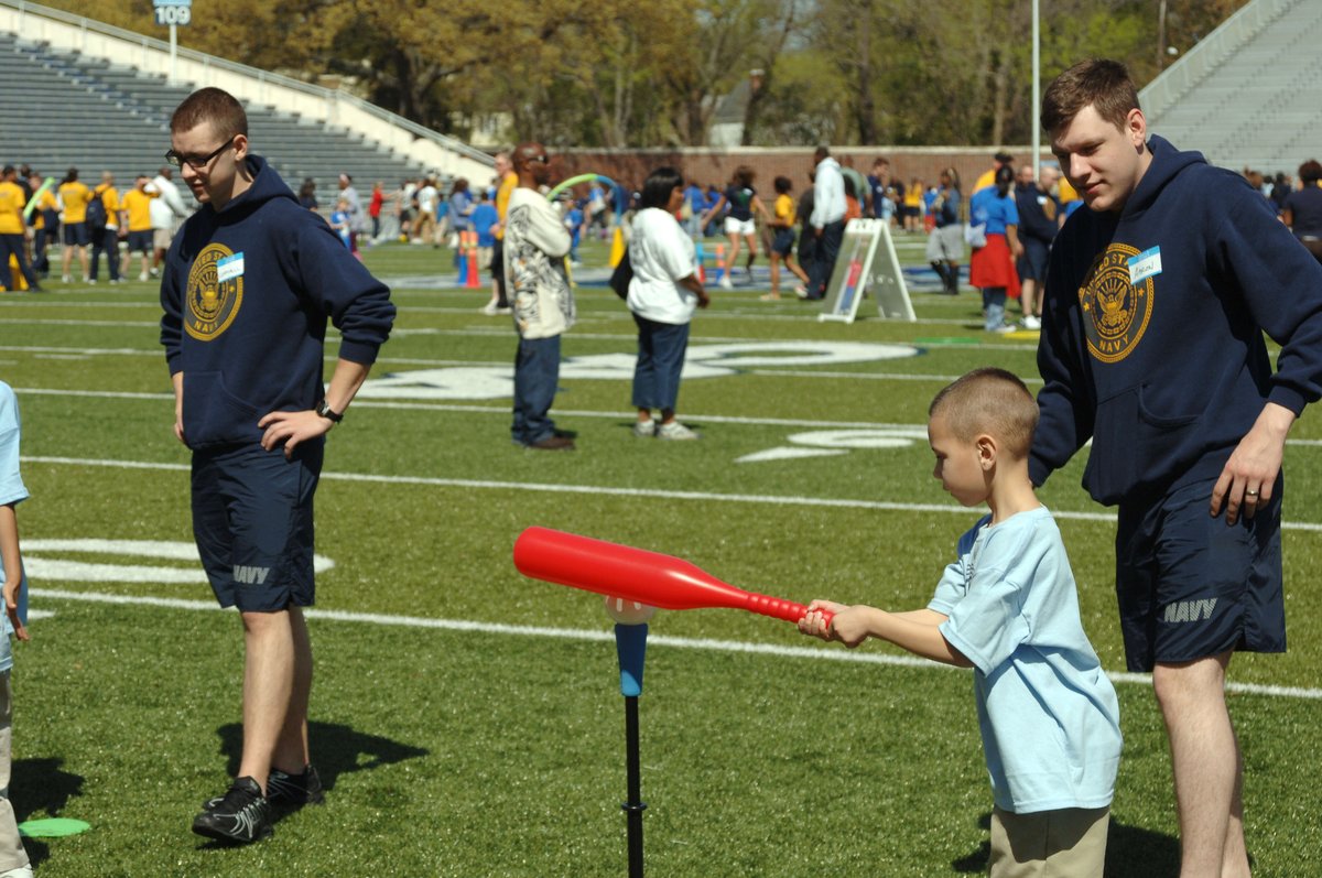 US Navy Operations Specialist 3rd Class Arron Wierckz helping a child hit a tee ball at Little Feet Meet