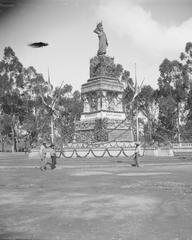 Monument to Quatemor in Mexico City featuring a statue with a feathered headpiece atop an ivy-covered stone monument surrounded by laurels, six flags on poles, and people walking by circa 1905-1910