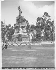 Monument to Quatemor in Mexico City circa 1905-1910 with statue of feathered headpiece figure atop ivy-covered stone monument surrounded by laurels and six flags flying on poles