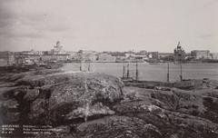 View from Tähtitorninmäki to Helsinki city center showing rocky foreground, sailboat masts, sea bay, and Helsinki waterfront with Tuomiokirkko and Uspenski Cathedral