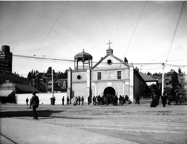 Nuestra Señora Reina de los Angeles Plaza Church with people gathered around, stone building with arched doorway and rooftop gazebo, historic Los Angeles
