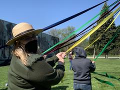 National Park Service ranger in straw flat hat and black KN95 mask holding yellow ribbon in May Pole dance