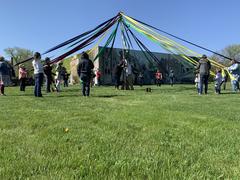 Community members of various ages participating in the May Pole Dance with colorful ribbons around a pole in a grassy field in Pullman during spring
