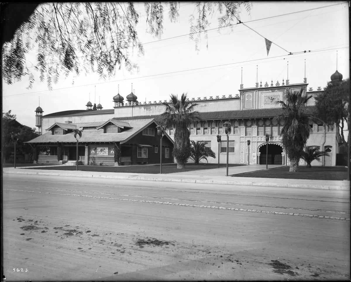 Exterior view of the First Shriners Auditorium in Los Angeles around 1910, showcasing Moorish-style architectural elements including Islamic horseshoe arches, ornamental moldings, battlement roofline, and onion-shaped towers. Lamp posts shaped like crescent moons line the pathway to the entrance, with a two-story house adjacent featuring stained glass windows with the Shriners emblem.