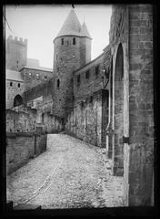View inside the medieval fortress of Carcassonne from the rampart, showing a path leading to a round turret with a pointed roof, a door on the right, and a square castle tower in the background