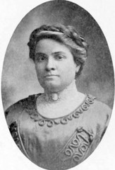 Portrait of a woman with an updo hairstyle wearing a high-collared satiny dress with a cameo and embroidered details