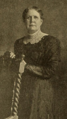 Portrait of Clara Marshall, an older white woman wearing a dark garment with a high white lace collar, standing with hands on a chair back