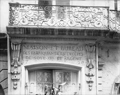 Detail of facade with wrought iron balcony and a sign reading Maison et bureau des fabricants en étoffes de soye or et argent 1727