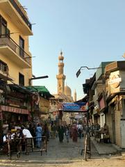 Al-Ashraf Mosque on busy Al-Muizz Street in Old Cairo, featuring intricate Mamluk-era geometric stonework on a colorful striped façade under a clear sky