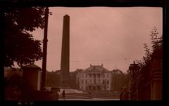 Obelisk memorial at Karolinenplatz in Munich with two men standing by a street lamp